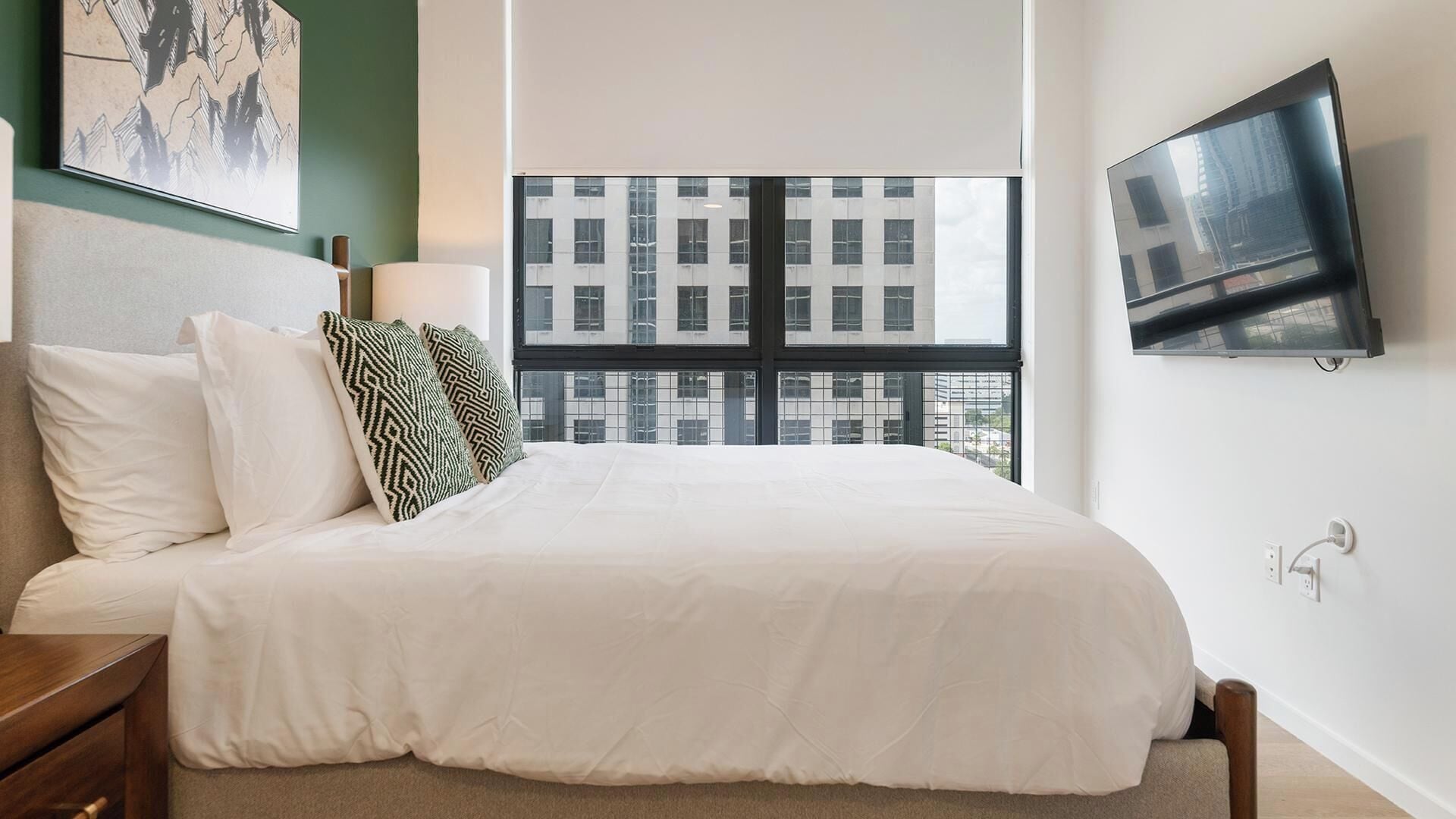 Ein Hotelzimmer mit einem großen Bett, einem an der Wand montierten Fernseher und einem Fenster mit Blick auf die Stadt.