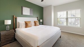 A hotel room with a large bed, two bedside tables, a nightstand, a window with blinds, and a green accent wall. - Landing | Spectacular 3BD, Gym (Fort Worth)