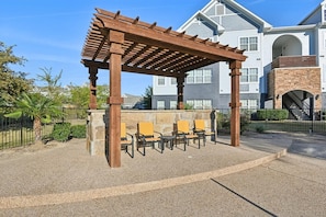 A wooden pergola with seating, a stone wall, and a building in the background. - Landing | Spectacular 3BD, Gym (Fort Worth)