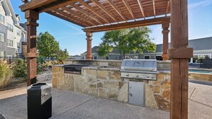 An outdoor barbecue area with a stone wall, a grill, and a pergola structure. - Landing | Spectacular 3BD, Gym (Fort Worth)