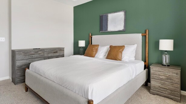 A neatly made bed with a wooden headboard, a grey dresser, a bedside table with a lamp, and a green accent wall. - Landing | Spectacular 3BD, Gym (Fort Worth)