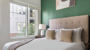 Ein Hotelzimmer mit einem großen Bett, einem Nachttisch, einer Lampe und Blick auf ein Gebäude.