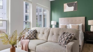 Ein modernes Wohnzimmer mit einem beigefarbenen Sofa, gemusterten Kissen, einer grünen Akzentwand und einem großen Fenster.