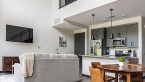 Eine moderne Küche mit Edelstahlgeräten, ein Essbereich mit Holztisch und -stühlen sowie ein Wohnbereich mit Sofa und Fernseher.