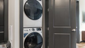 Ein Waschraum mit einer Frontlader-Waschmaschine und einem Trockner, einem Metallmülleimer und einer Tür zu einem weiteren Raum.