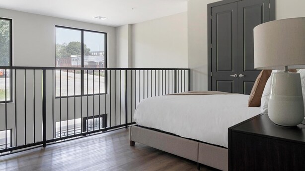 A bedroom with a large bed, a nightstand, a lamp, a wardrobe, and a balcony with a railing. - Landing | Spectacular 1BD, Gym, Courtyard (Atlanta)