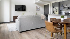 Ein modernes Wohnzimmer mit grauem Sofa, einem Esstisch mit Stühlen, einer Küchenzeile mit Geräten und einem an der Wand befestigten Flachbildfernseher.
