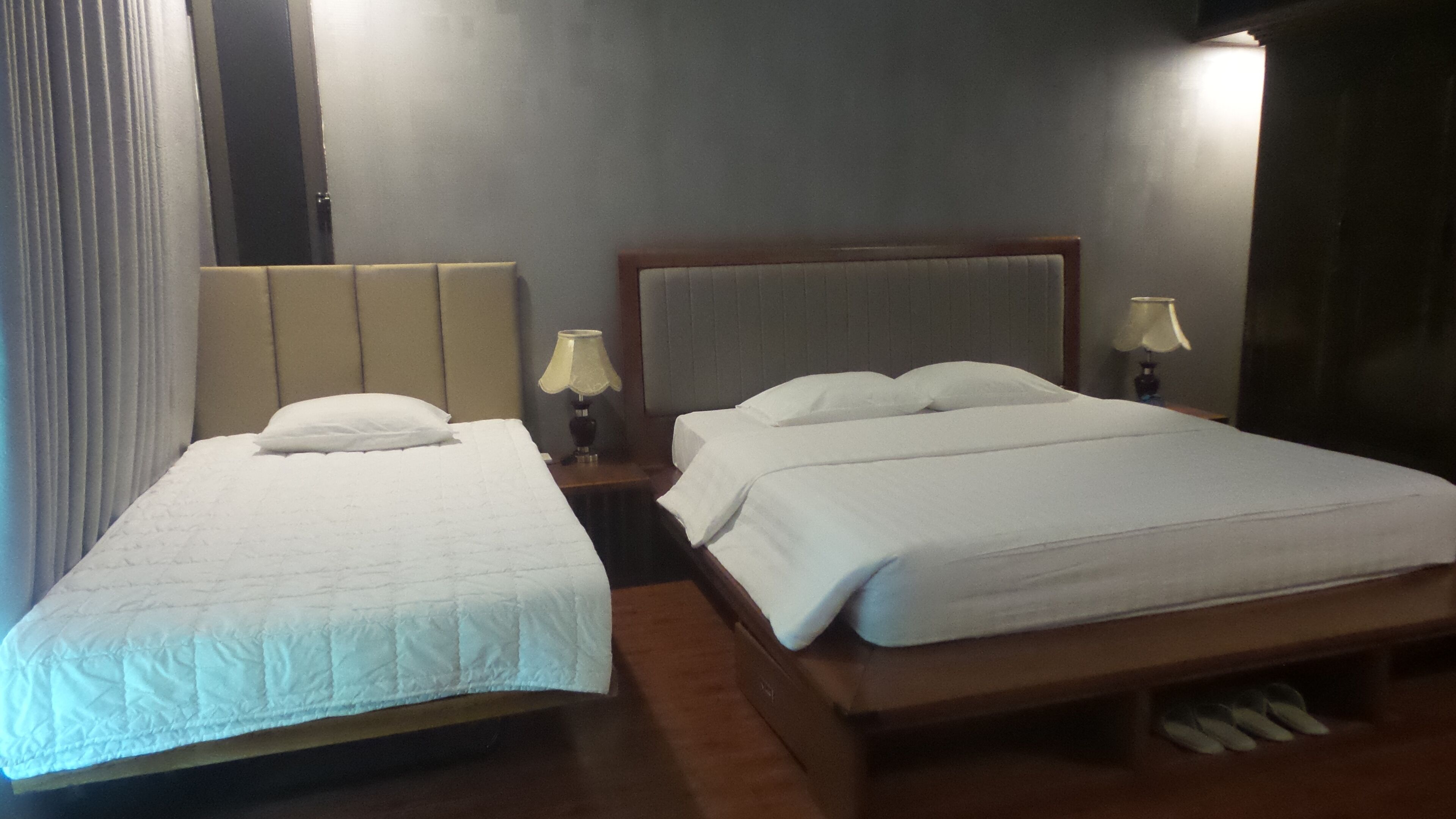 A hotel room with two beds, a wooden headboard, a nightstand with a lamp, and a window with curtains.