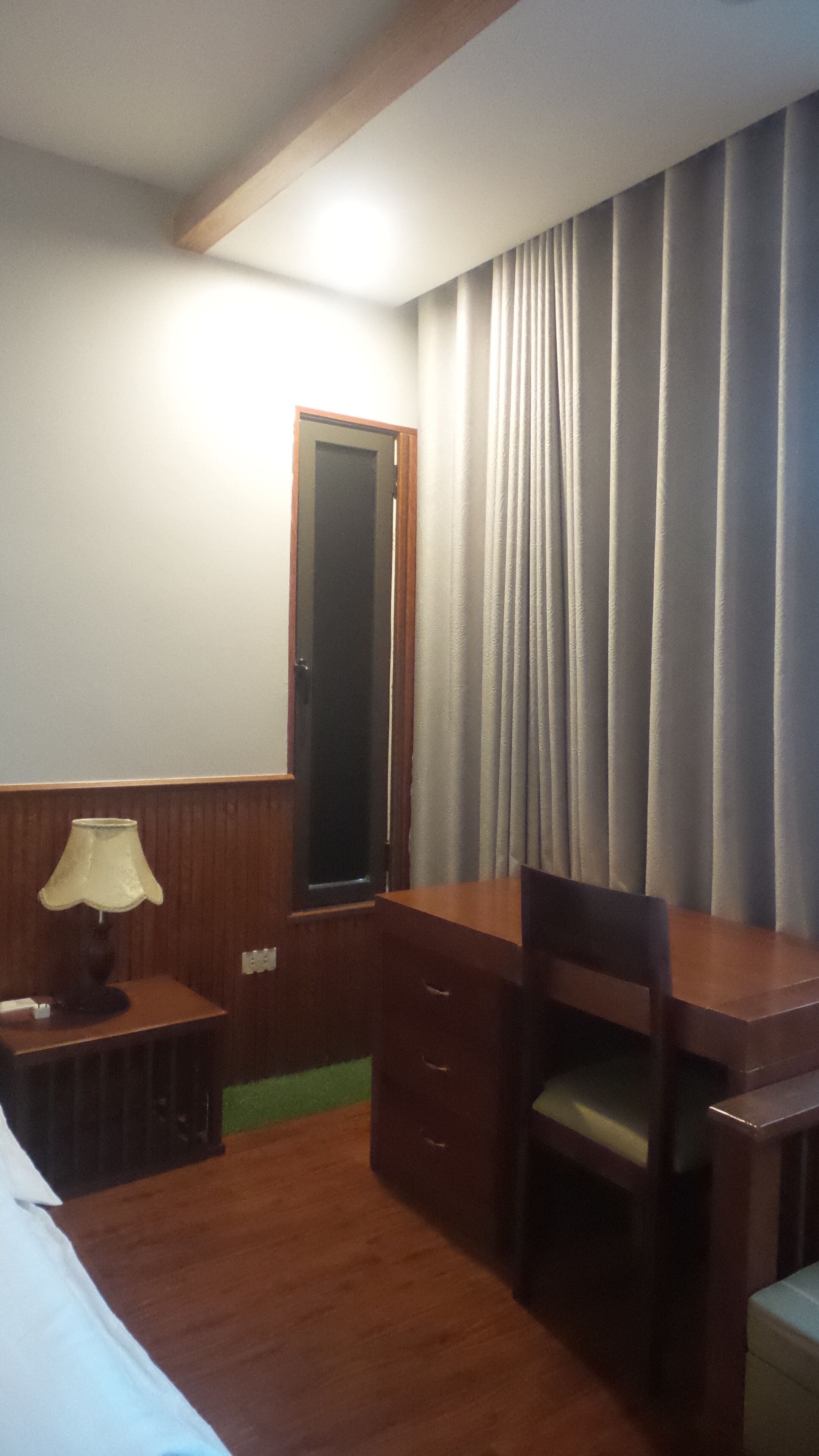 Quarto de hotel com escrivaninha de madeira, uma cadeira, abajur, espelho e janela com cortinas.