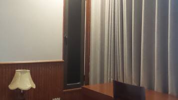 Quarto de hotel com escrivaninha de madeira, uma cadeira, abajur, espelho e janela com cortinas.