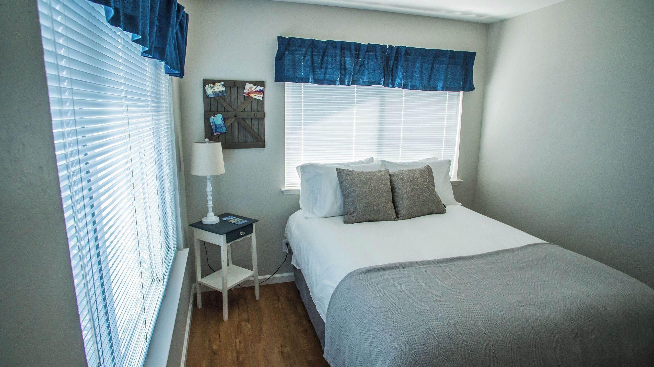 A bedroom with a bed, a nightstand, a lamp, a window with blinds, and a wall-mounted picture.