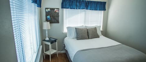 Un dormitorio con cama, mesita de noche, lámpara, ventana con persianas y un cuadro colgado en la pared.