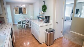 A kitchen with white cabinetry, a dining table with chairs, a coffee maker, and a trash can. - Vintage Paso by Avantstay Firepit + Garden + BBQ Walk to Downtown Paso (Paso Robles)