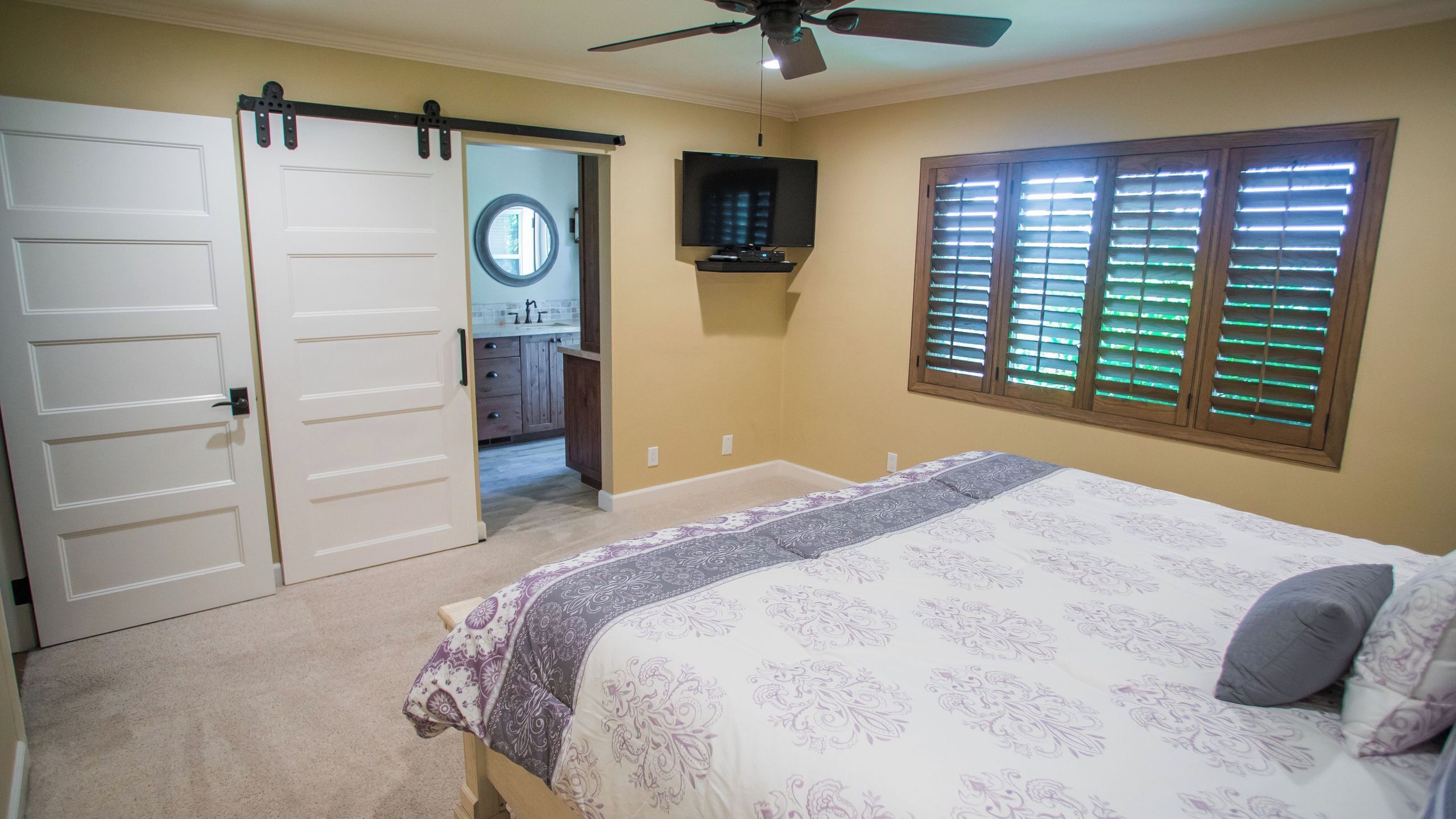 Quarto com cama, ventilador de teto, TV e janela com persianas.