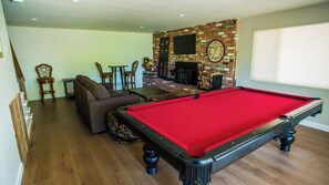 Ein Billardtisch in einem Raum mit Kamin aus Ziegelsteinen, einer Couch und einem an der Wand befestigten Fernseher.