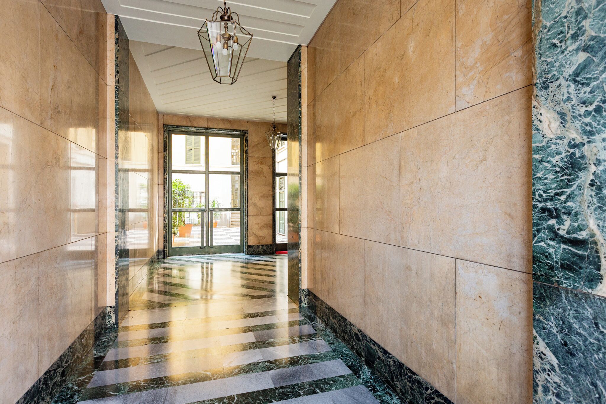 Um corredor com piso xadrez, paredes de mármore e um lustre.
