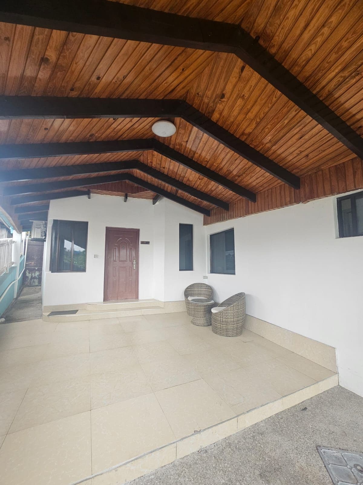 Un espace extérieur abrité, doté d’un plafond en bois, d’un mur blanc avec une porte et de deux fauteuils en osier.