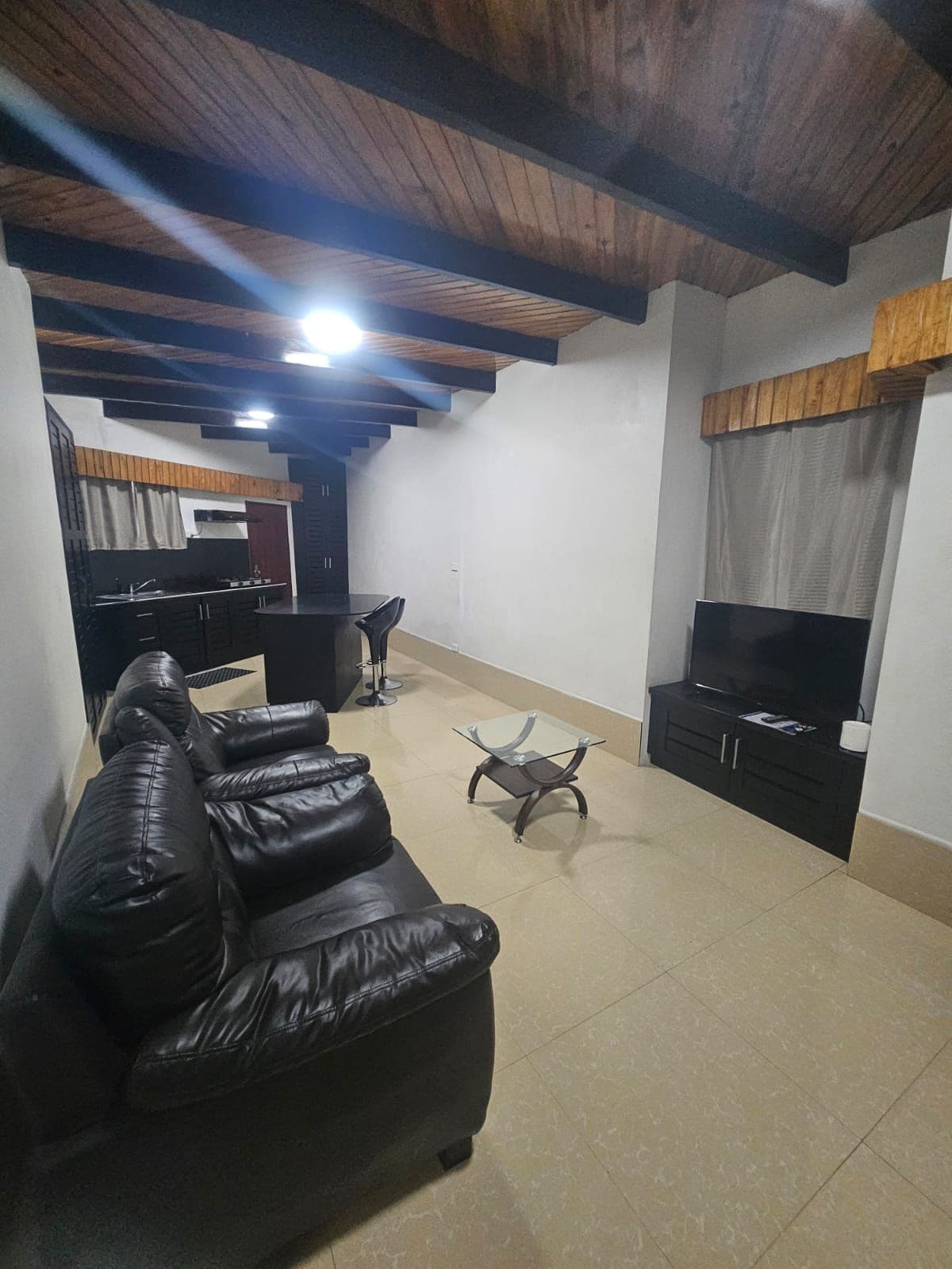 A living room with a black leather sofa, a glass coffee table, a TV stand, and a wooden ceiling.