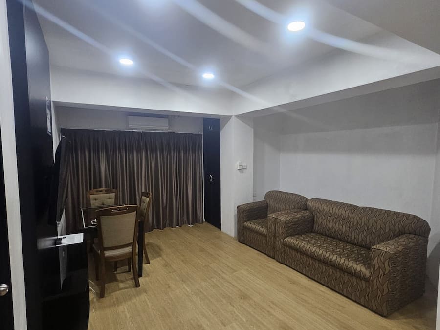 Um quarto com piso de madeira, um sofá marrom com estampa, uma pequena mesa com uma cadeira e um armário preto.