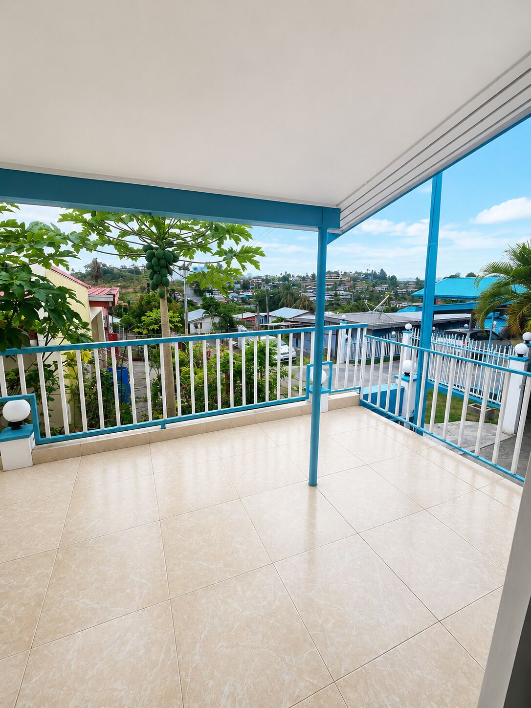 Varanda com chão de azulejos, guarda-corpo azul e vista para uma área residencial.