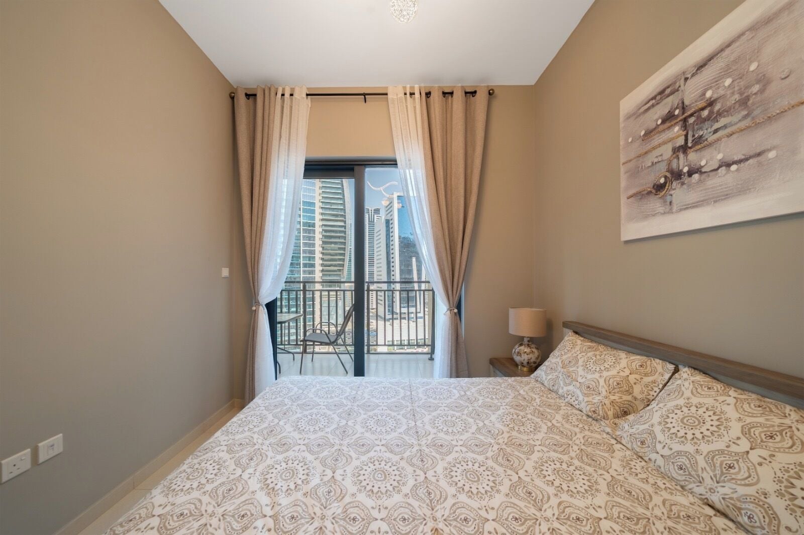 A hotel room with a bed, a nightstand, a lamp, a chair, and a balcony with a view of tall buildings.