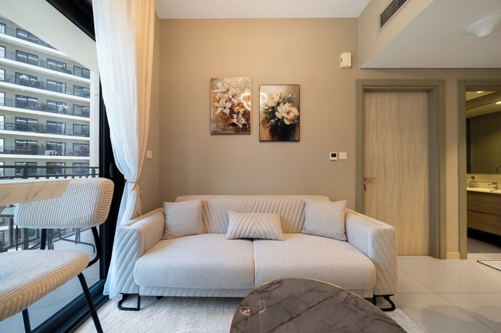 Ein modernes Wohnzimmer mit einer Couch, einem kleinen Tisch und einem Sessel. An der Wand hängen zwei Bilder, eine Tür ist zu sehen und es gibt einen Blick auf ein Gebäude.