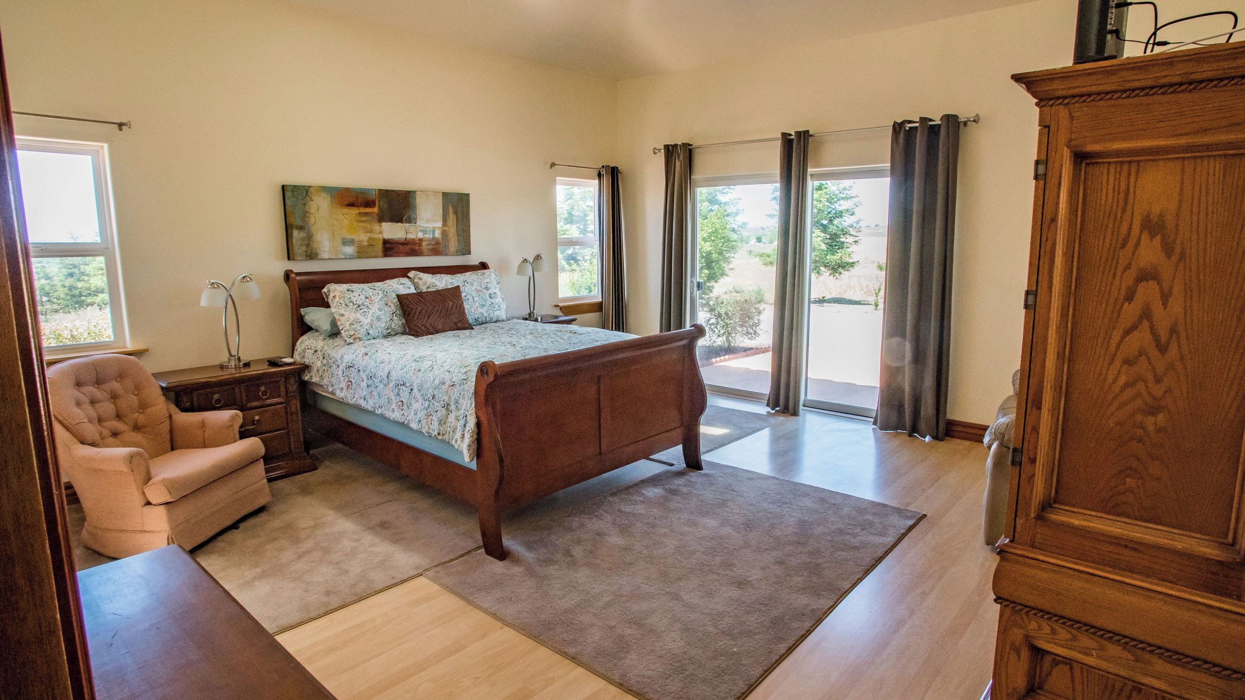 A bedroom with a large bed, a chair, a nightstand, a wooden cabinet, and a view of the outdoors through the window.