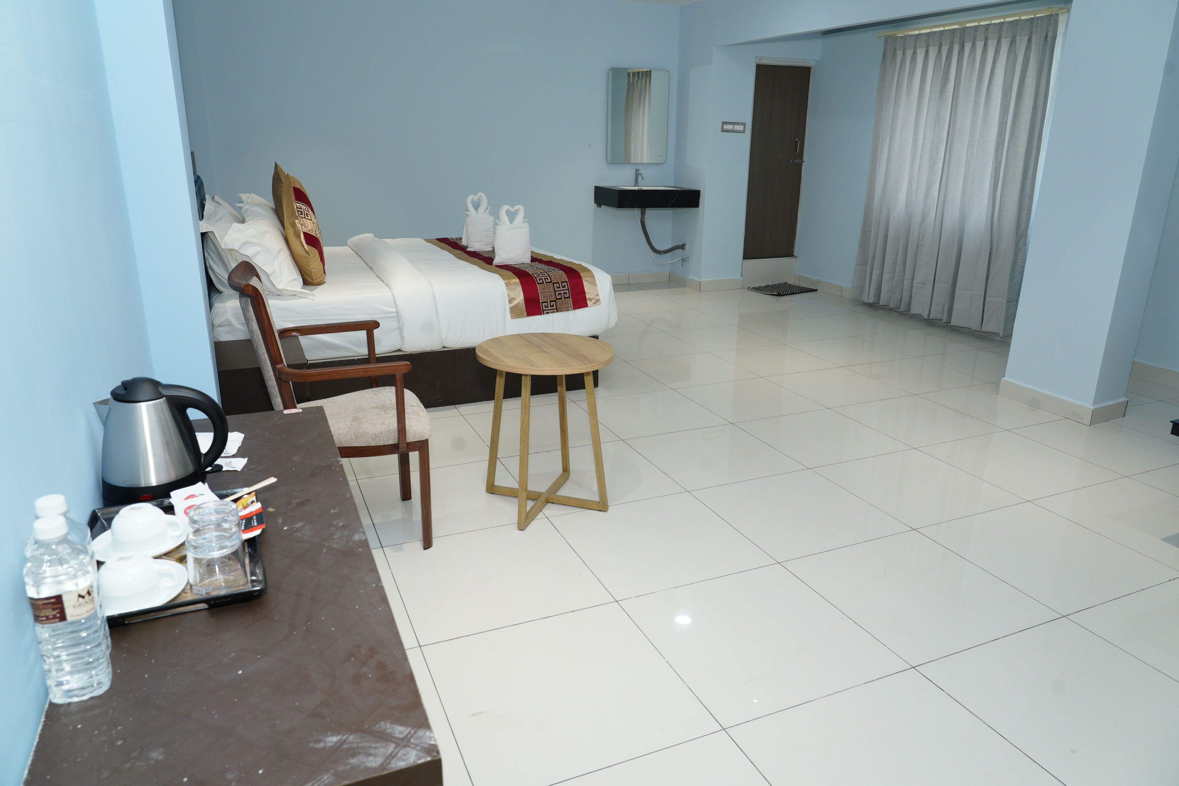 A hotel room with a bed, a small table with a kettle, a chair, and a bedside table with a water bottle and glasses.
