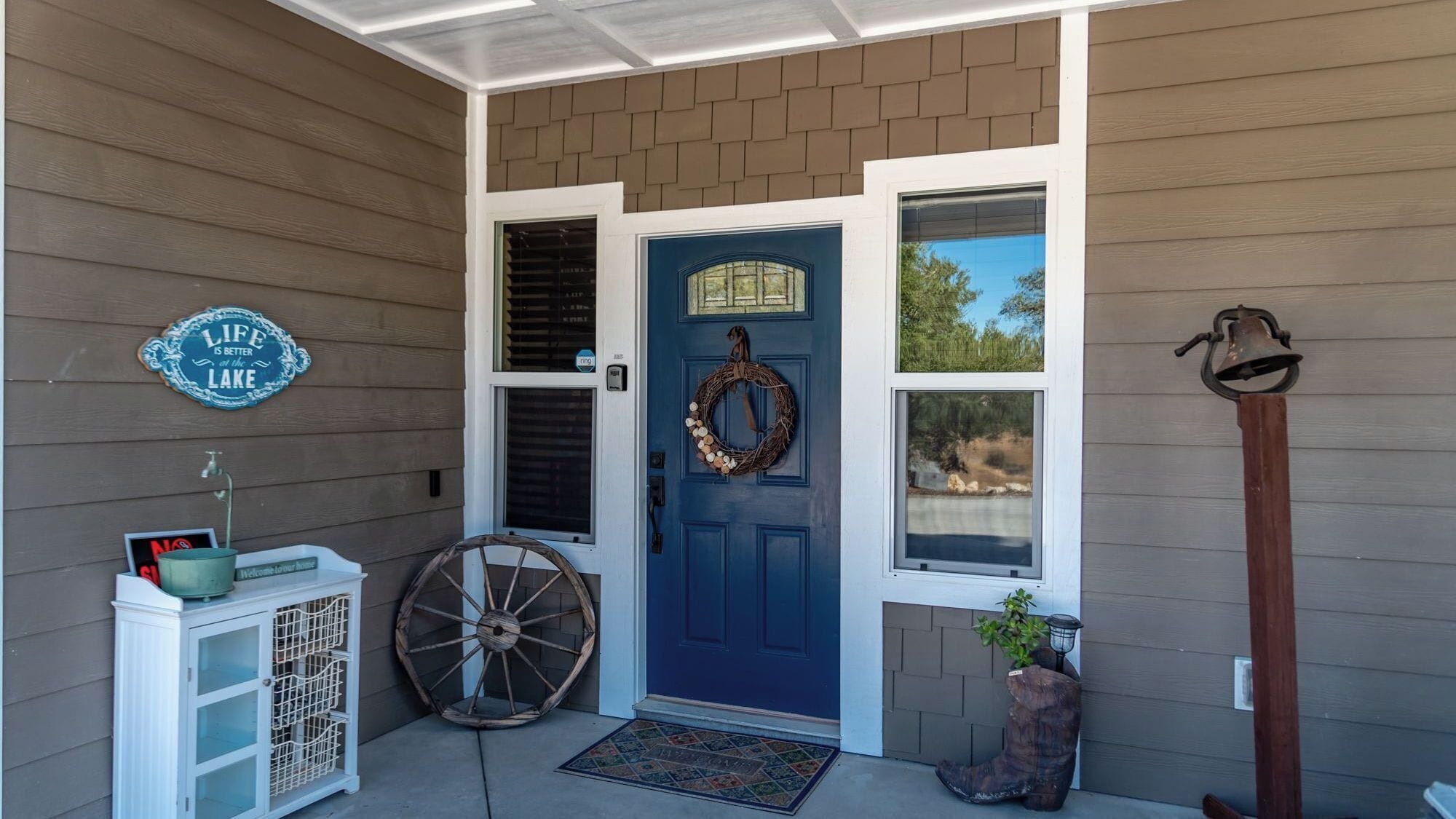 A front porch with a blue door, a welcome sign, a wreath, a wooden wheel, a cabinet, and a bell on a post.