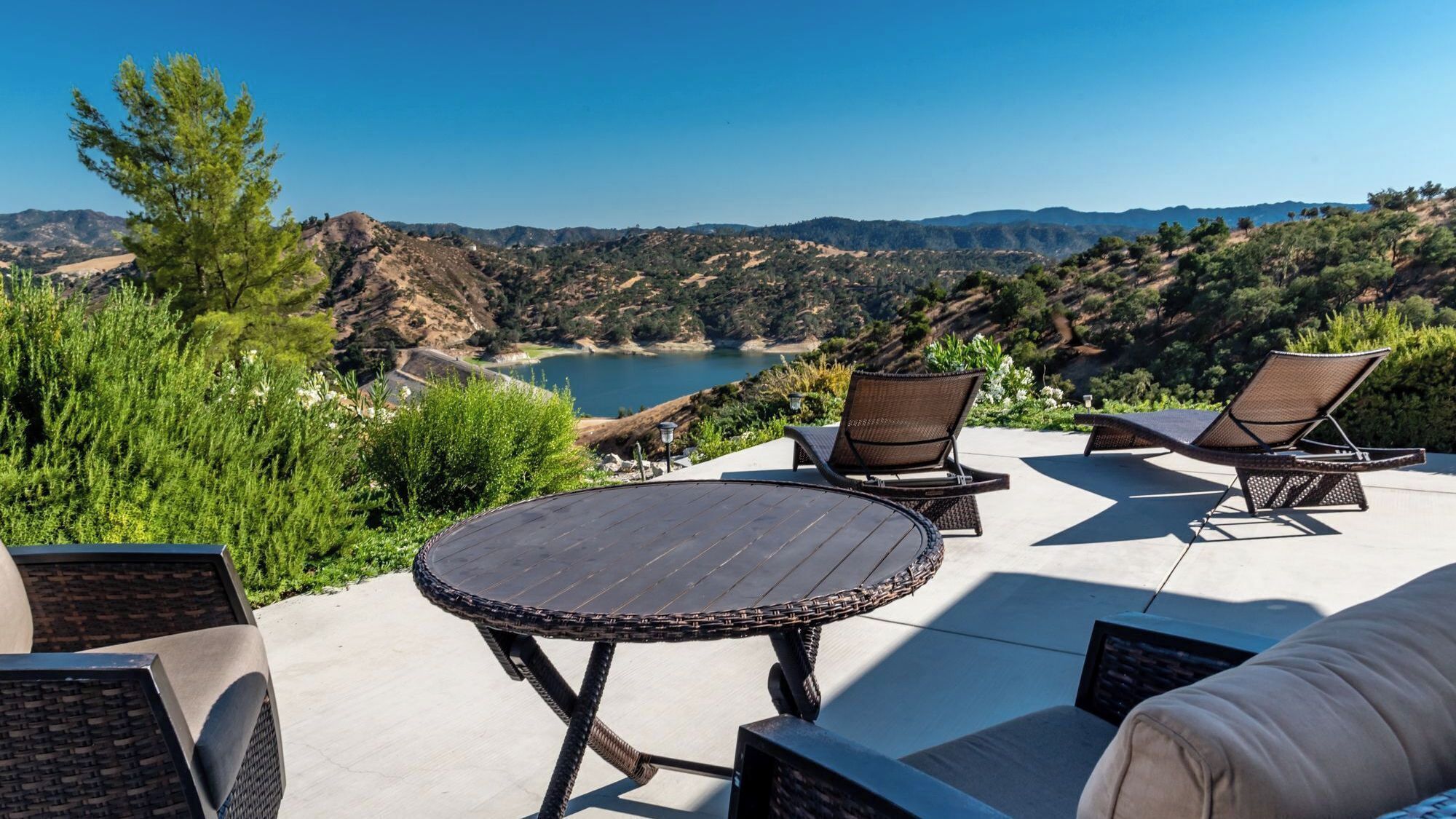 An outdoor patio with wicker furniture, a round table, and a view of a lake and mountains.