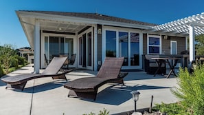 Une terrasse extérieure couverte, dotée de deux chaises longues et d’une table, ainsi qu’une pergola et une maison avec de grandes fenêtres.