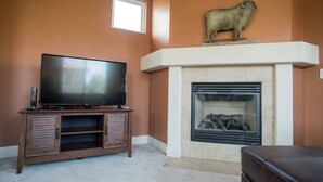 A living room with a flat-screen TV on a wooden cabinet, a fireplace with a stone mantle, and a stone sculpture of a bull on the wall. - Navajo Trail by Avantstay Lap Pool + Spa Steps to Salinas River Trail (Paso Robles)