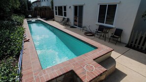 Ein rechteckiger Pool mit Fliesenrand, umgeben von einer Terrassenfläche mit Stühlen und einem Tisch.