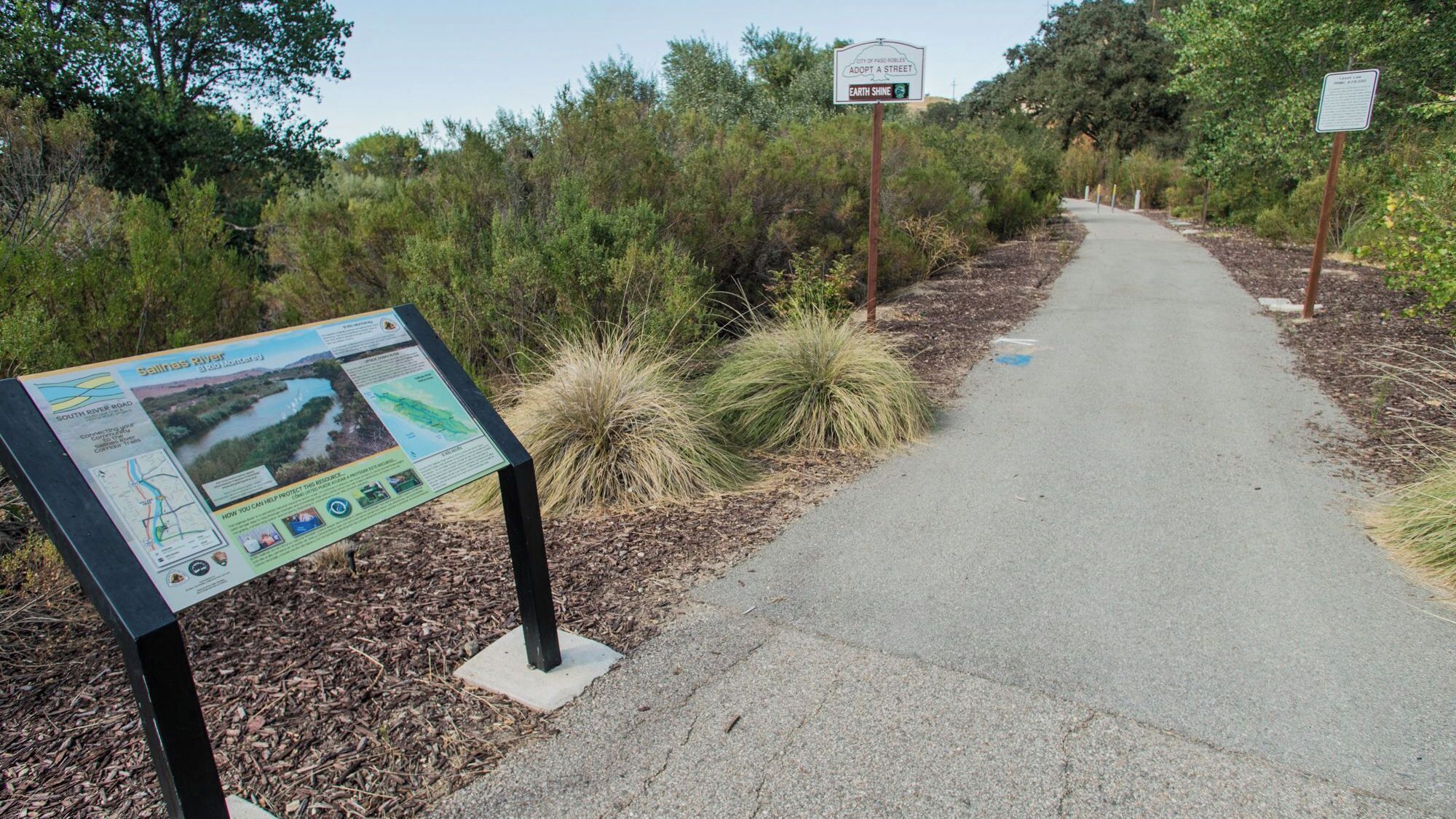 A paved path with informational signs, surrounded by vegetation.