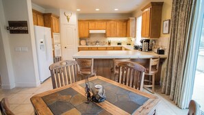 A kitchen with wooden cabinets, a white refrigerator, a central island with bar stools, and a dining table with a decorative centerpiece. - Navajo Trail by Avantstay Lap Pool + Spa Steps to Salinas River Trail (Paso Robles)
