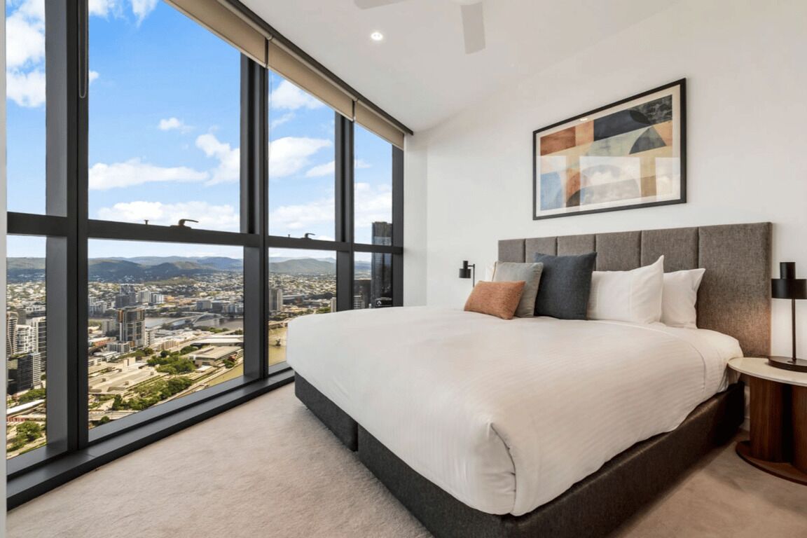 Un dormitorio con una cama grande, una mesita de noche, una lámpara y vistas de la ciudad a través de amplias ventanas.