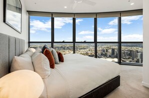 Une chambre à coucher avec un grand lit, de nombreux oreillers, une vue sur le paysage urbain à travers de grandes fenêtres et un ventilateur de plafond.