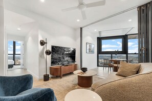 Un salon moderne avec un canapé, une table basse et un téléviseur. Une grande fenêtre offre une vue sur la ville.