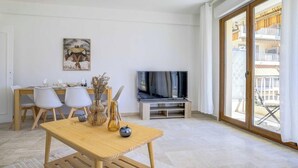 Una sala de estar moderna con una mesa de madera, sillas blancas, un televisor de pantalla plana y un balcón con mobiliario exterior.