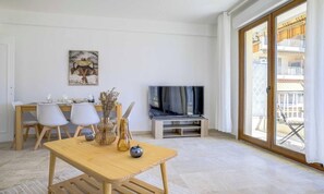 Ein modernes Wohnzimmer mit Holztisch, weißen Stühlen, einem Flachbildfernseher und einem Balkon mit Gartenmöbeln.