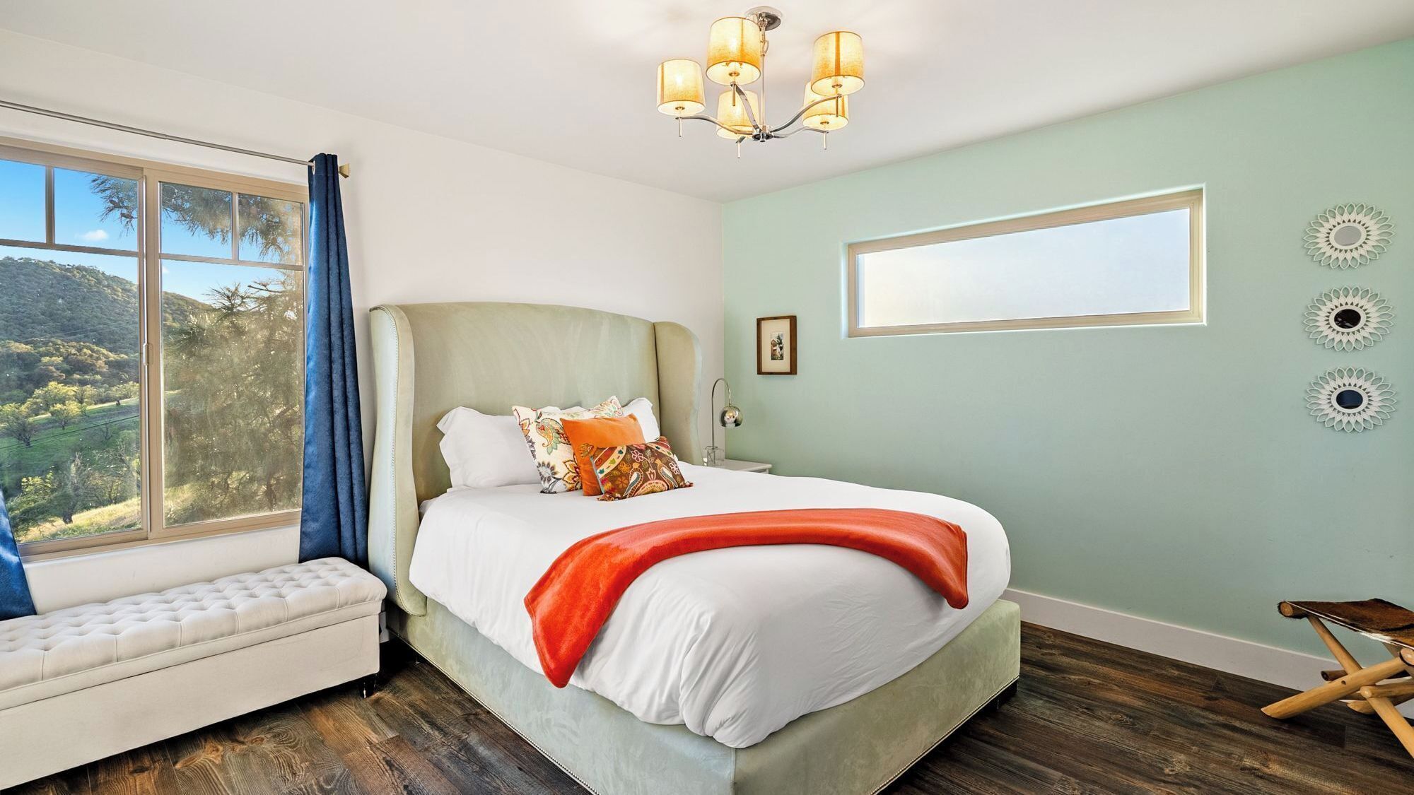 A bedroom with a large bed, a window with a view of the outdoors, a chandelier, and a bedside table.