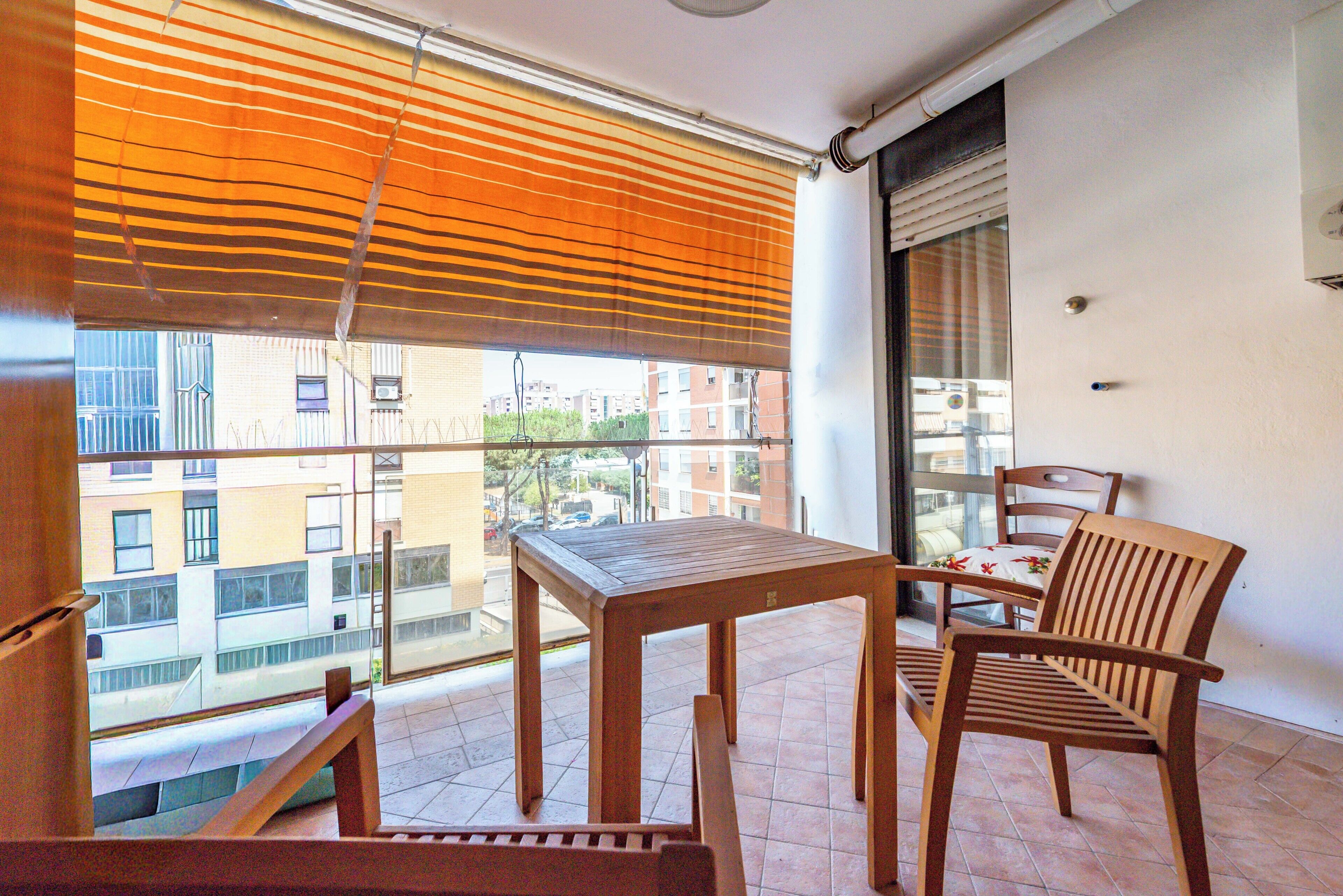 A balcony with a wooden table and two chairs, a window with blinds, and a view of buildings.