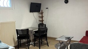Ett litet, väl upplyst rum med en tv monterad på väggen, ett matbord med stolar, en röd soffa och en liten matta på golvet.