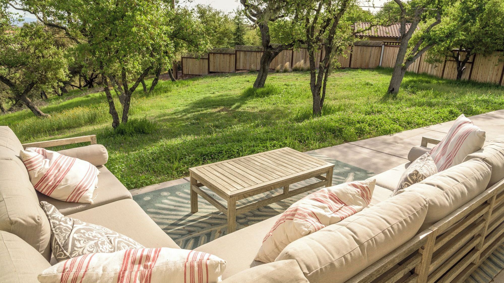 An outdoor seating area with a wooden table and cushioned sofas, surrounded by a wooden fence and trees.