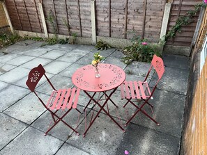 Une terrasse avec une table ronde et deux chaises, un petit vase avec des fleurs et, au fond, une clôture en bois.