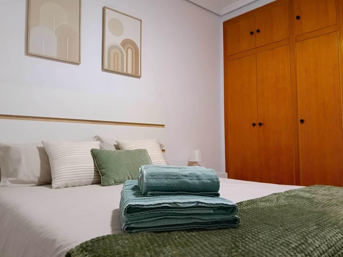 Un lit bien fait, avec des oreillers et une serviette pliée, une armoire en bois et deux œuvres d’art encadrées accrochées au mur.