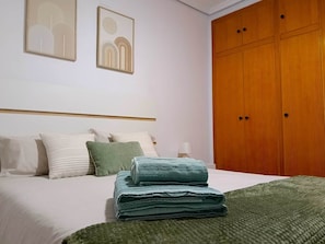 Un lit bien fait, avec des oreillers et une serviette pliée, une armoire en bois et deux œuvres d’art encadrées accrochées au mur.