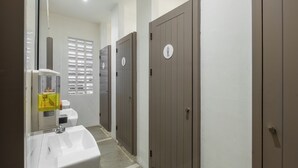 A bathroom with a sink, toilet, and three doors, each with a gender symbol. - New Hostel Braga (Bandung)