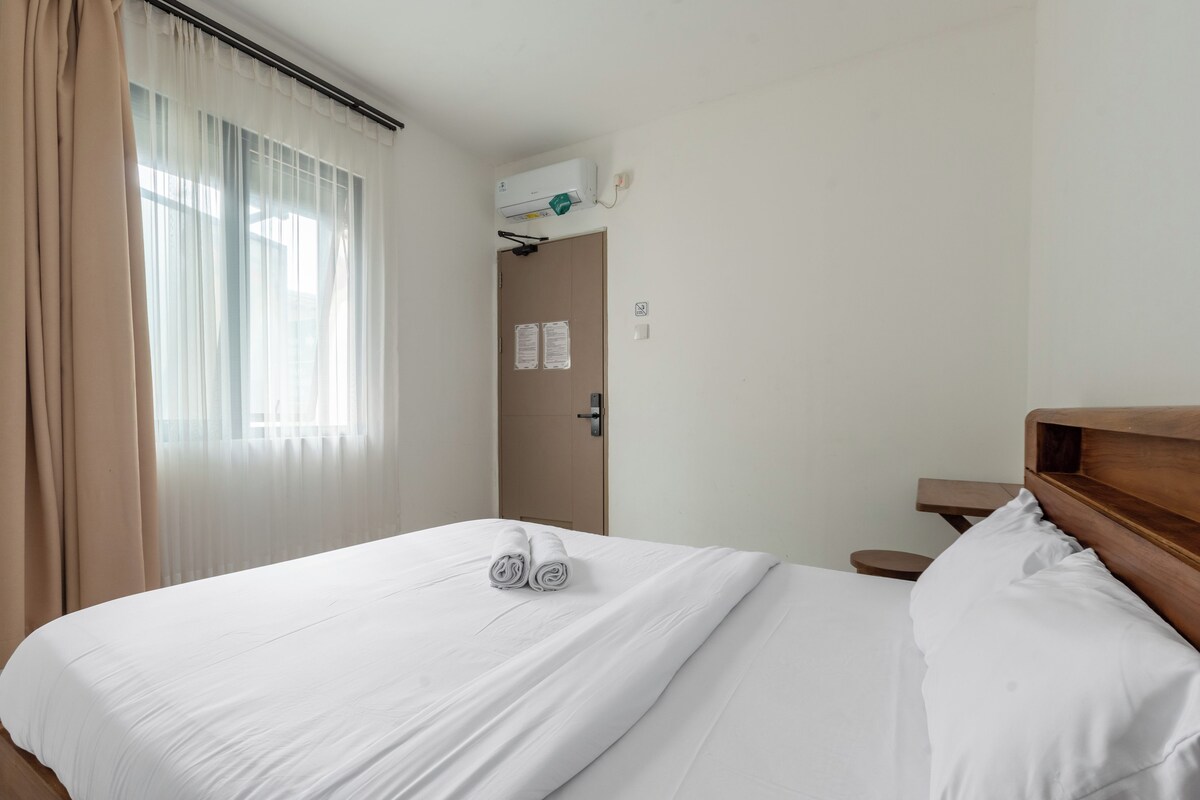 A hotel room with a bed, a wooden headboard, a nightstand, a window with sheer curtains, and a door with a light switch.