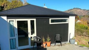 A small building with a dark roof, a sliding glass door, and a patio area with two chairs and potted plants. - Contempory Hillside studio in Cashmere (Christchurch)